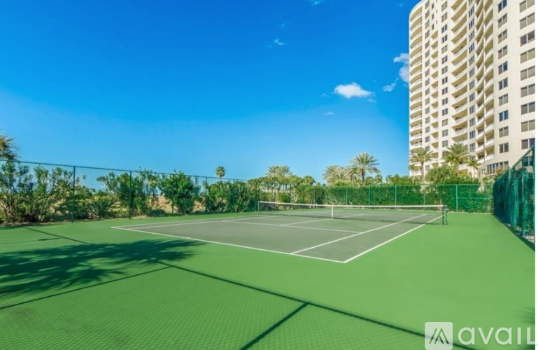 A tennis court surrounded by a fence and trees with a building in the background.