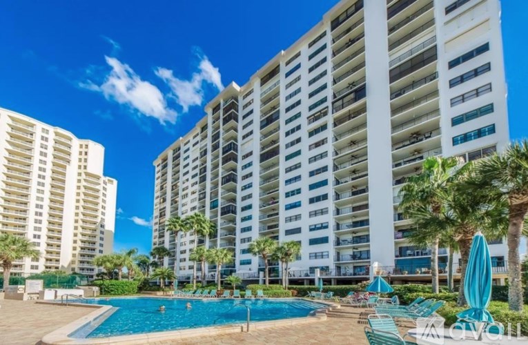 A large swimming pool is surrounded by lounge chairs and umbrellas in front of a large apartment building.