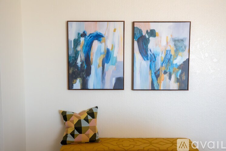 Two abstract paintings hang on a wall above a yellow couch with a multicolored pillow.
