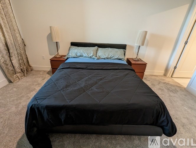 A bed with a black comforter and two pillows sits in a room with carpeted flooring and two lamps on wooden nightstands.