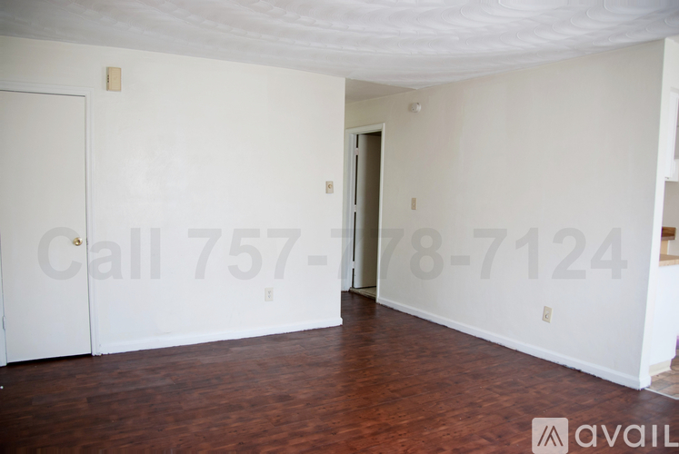 A room with a wooden floor and white walls, with a door on the left and a partially visible door on the right.