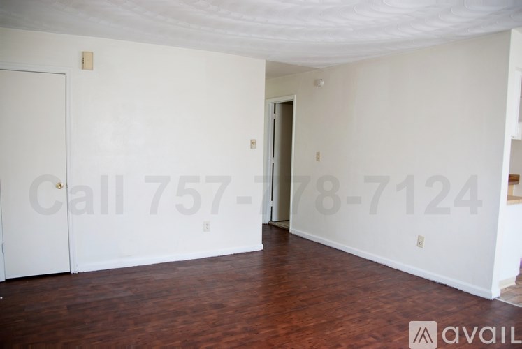 A room with a wooden floor and white walls, with a door on the left and a partially visible door on the right.