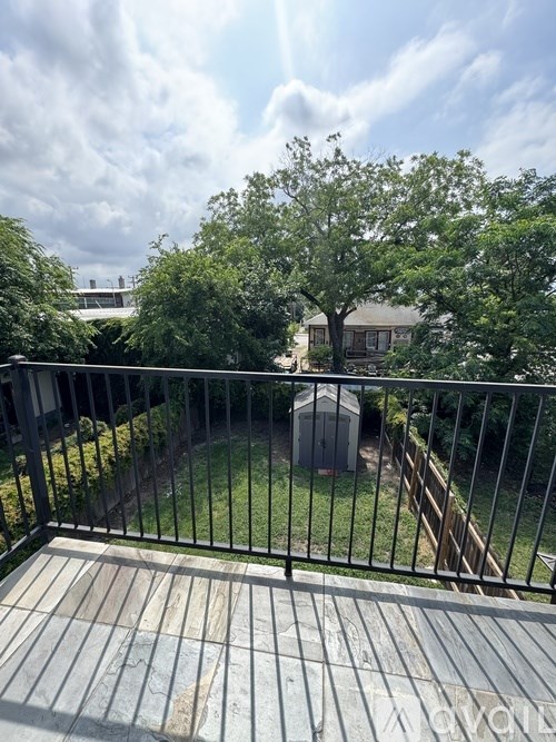 A balcony with a metal railing and a concrete floor overlooks a backyard with a tree and a shed.