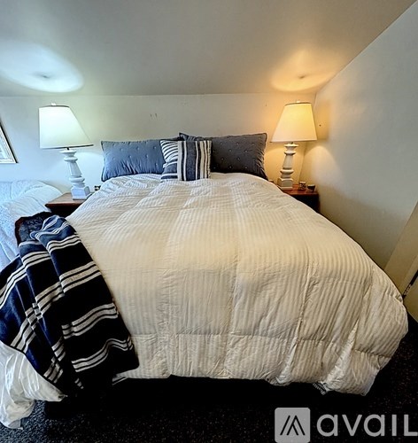 A bed with a white comforter and a striped blanket is in the foreground of a room with two lamps on bedside tables.