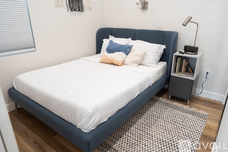 A bed with a white sheet and a blue headboard is in a room with a lamp and a bookshelf.