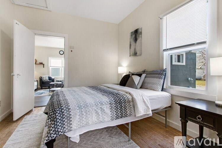 A bedroom with a bed covered in a patterned blanket and pillows, a nightstand, and a window with blinds.