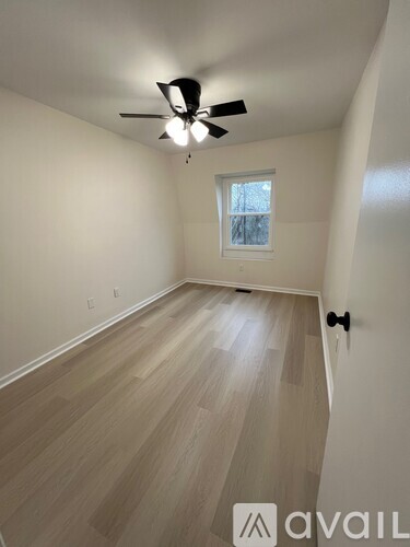 A room with a ceiling fan and light, wooden flooring, and a window with a view of trees outside.