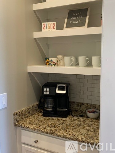 A coffee maker is on a countertop with a sign that says "More coffee please!" and a number "27502" on a shelf above it.