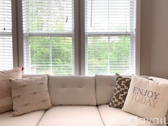 A white couch with pillows and a pillow that says "enjoy today" in front of a window.