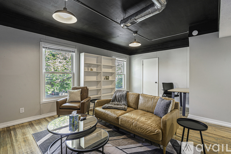 A living room with a brown leather couch, a glass coffee table, and a window with a view of trees.