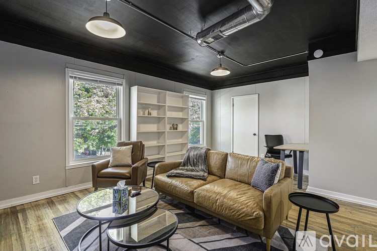 A living room with a brown leather couch, a glass coffee table, and a window with a view of trees.