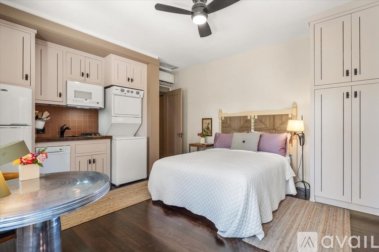A bedroom with a bed, a table, and a kitchen area with appliances.