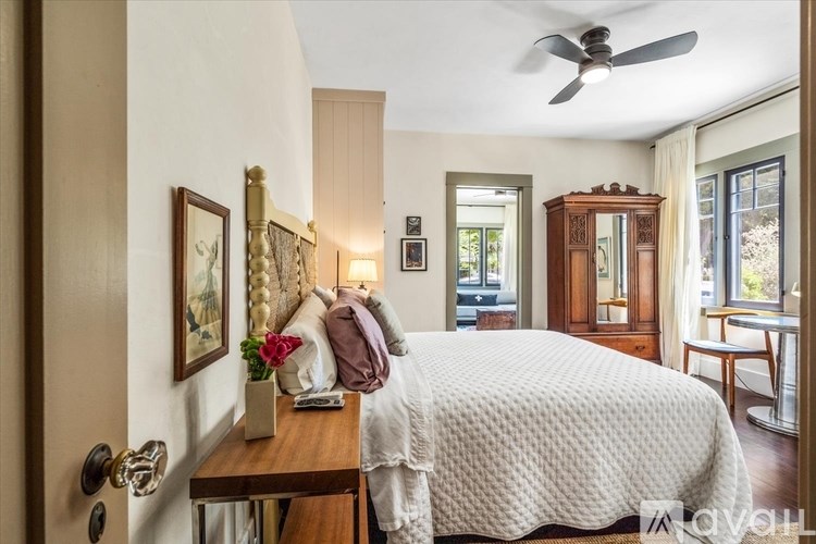 A bedroom with a bed, a dresser, a nightstand, a chair, a table, a vase, a fan, a window, a door, a painting, a lamp, a wall, a curtain, a wall,