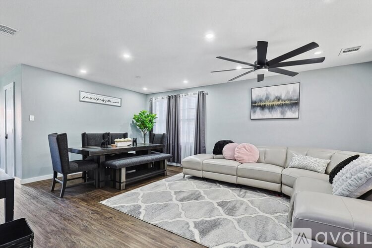 A living room with a grey couch, a black chair, a dining table with chairs, a rug, a ceiling fan, and a painting on the wall.