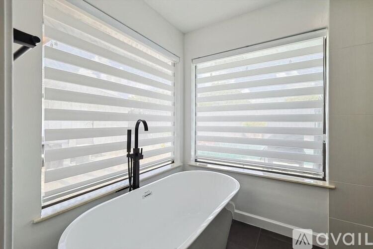 A white bathtub sits in a bathroom with a window covered in white blinds.