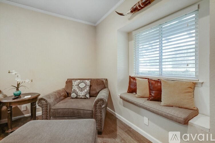 A living room with a brown couch, a grey ottoman, and a small table with a vase of flowers on it.