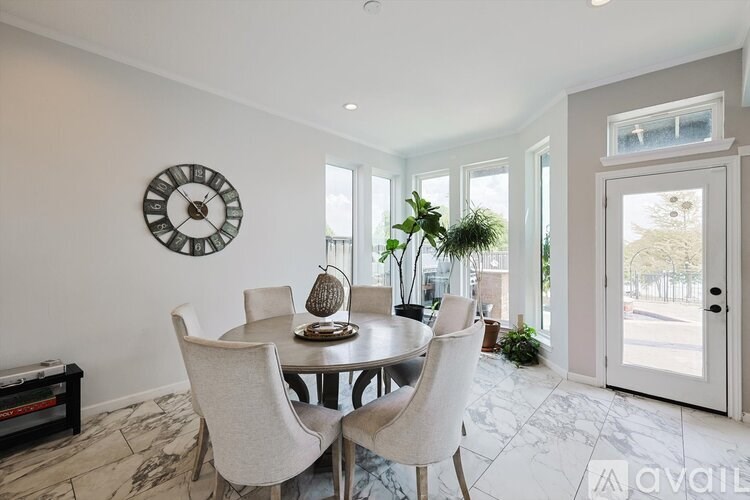A dining room with a round table and chairs, a clock on the wall, and a view of the outside through the glass doors.