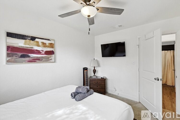 A bedroom with a bed, a ceiling fan, a nightstand with a lamp, a painting on the wall, and a flat-screen TV mounted on the wall.