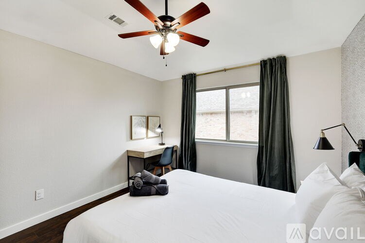 A bedroom with a large bed, a desk with a chair, a window with curtains, and a ceiling fan.