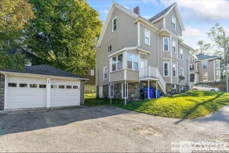 A two-story residential building with a garage is surrounded by a gravel driveway and trees.