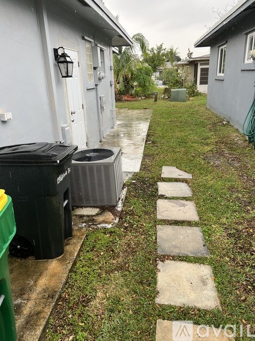 A wet walkway leads to a house with a green trash can on the left.