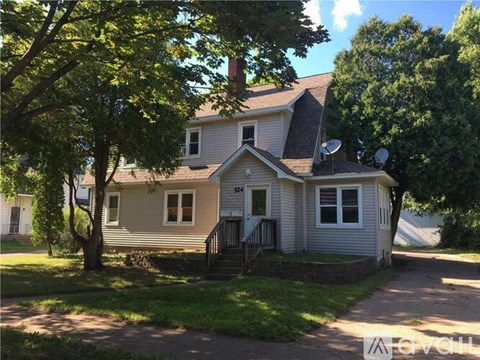 A house with a grey siding and a white door is surrounded by green trees.