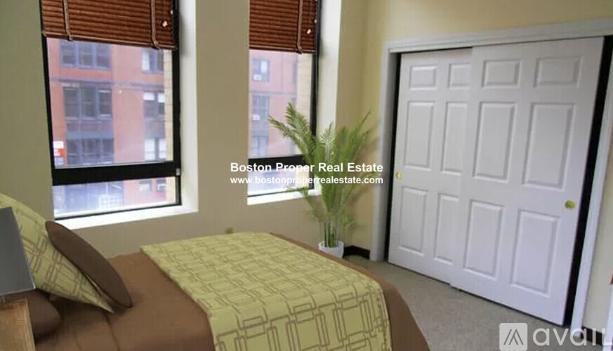 A bedroom with a bed, pillows, a window with blinds, a potted plant, and a white door.