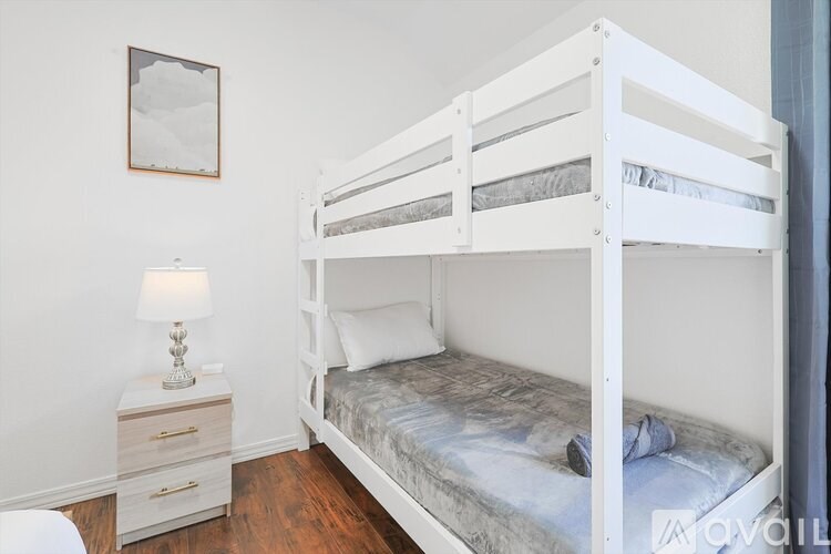 A white bunk bed with a grey mattress and a white nightstand with a lamp on it.