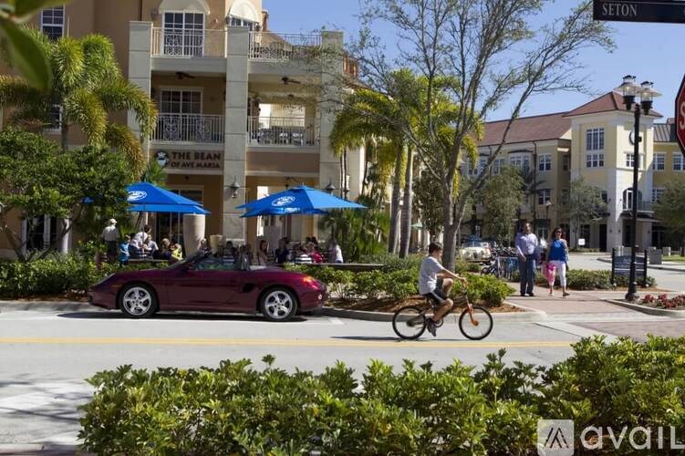 A man rides a bicycle down a street lined with palm trees and a red convertible car is parked on the side of the road.