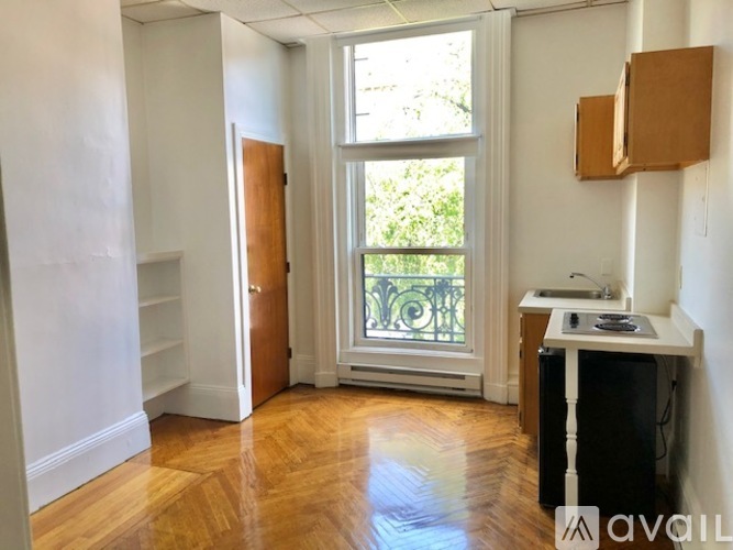 A room with a wooden floor, a window with a view of greenery outside, and a desk with a computer.