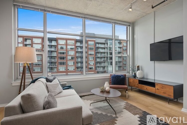 A living room with a grey couch, a round table, a wooden cabinet, and a large window overlooking a building.