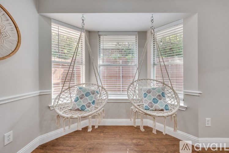 Two white hanging chairs with blue cushions are suspended from a window with blinds.