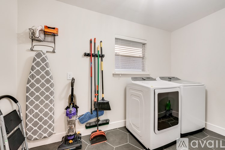 A white room with a vacuum cleaner, a broom, a mop, a baby stroller, and a couple of bottles in a mini fridge.