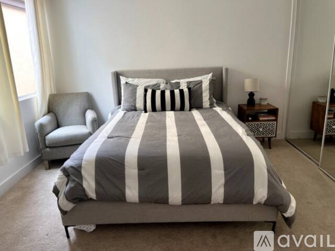 A bed with a grey and white striped comforter and two striped pillows sits in a room with a grey chair and a small table with a lamp.