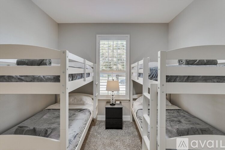 A room with white walls and a carpeted floor has two bunk beds and a small table with a lamp on it.