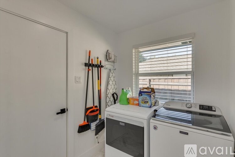 A white washing machine is in a small laundry room with a white door and a window with blinds.