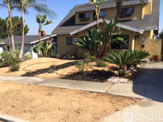 A house with a brown roof and a yellow wall is surrounded by plants and trees.