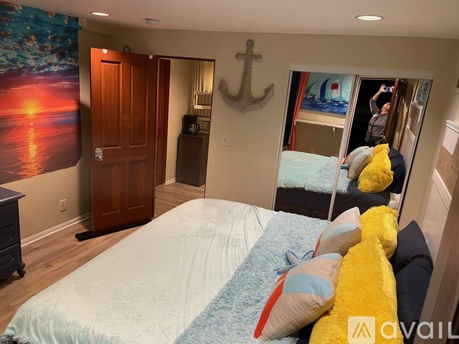A bedroom with a bed covered in pillows and a painting of a sunset on the wall.