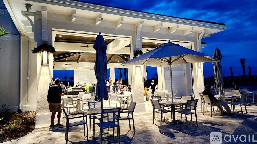 A restaurant with tables and chairs under umbrellas with people standing and sitting.