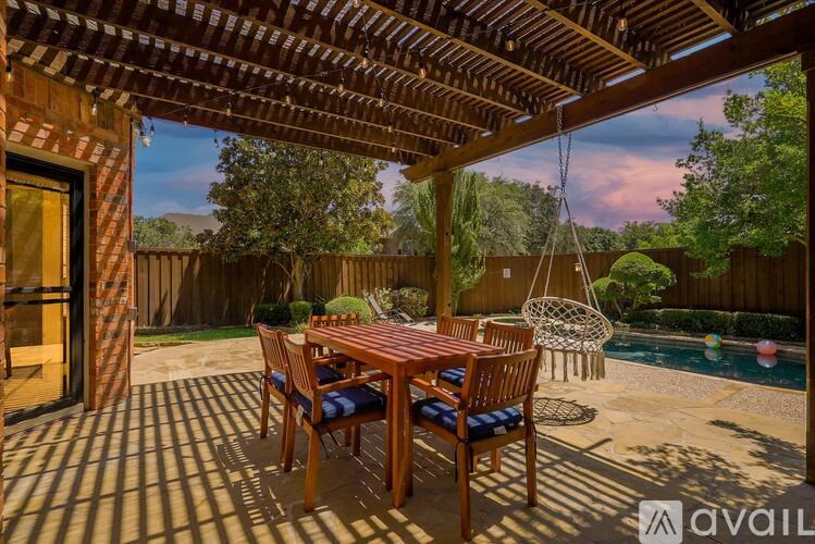 A wooden table and chairs are set up on a patio with a hanging chair and a pool in the background.