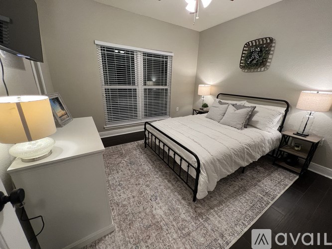 A bedroom with a large bed, a nightstand with a lamp, and a window with blinds.