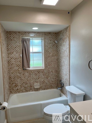 A bathroom with a white tub and toilet, a window with a brown curtain, and a tiled wall.