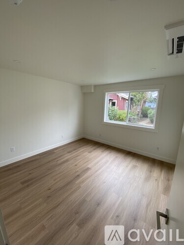 A room with wooden flooring and a window showing a view of a red building and trees.