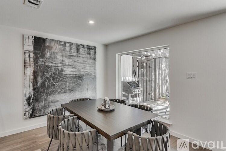 A dining room with a wooden table and chairs, a large black and white painting on the wall, and a view of a kitchen through a glass door.