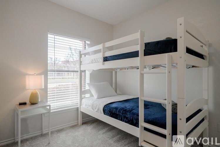 A white bunk bed with a blue mattress is in a room with a white nightstand and a lamp.