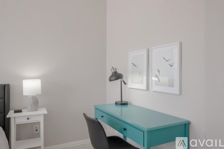 A turquoise desk with a black chair and a white nightstand with a lamp.