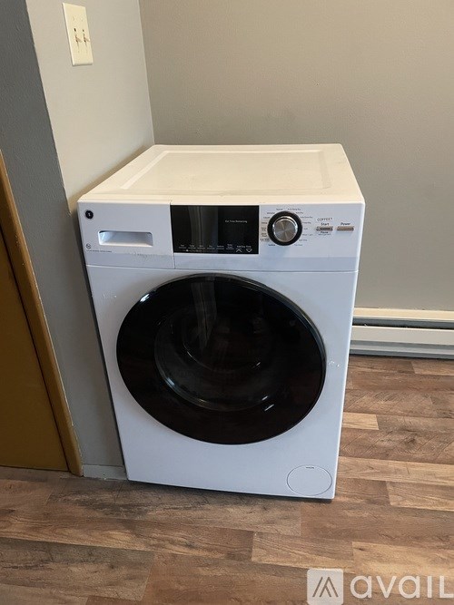 A white front loading washing machine with a black door is placed on a wooden floor.