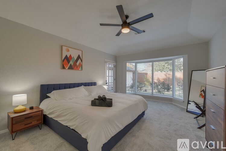 A bedroom with a large bed, a ceiling fan, and a view of the backyard through the windows.