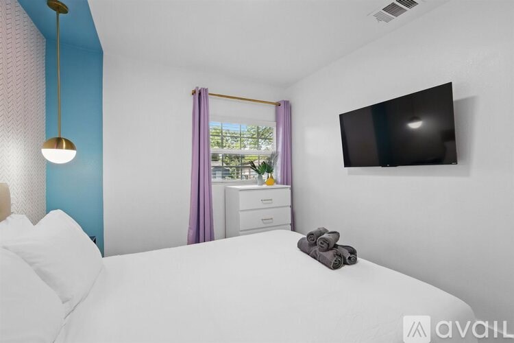 A bedroom with a bed, a dresser, a window with curtains, and a flat-screen TV mounted on the wall.