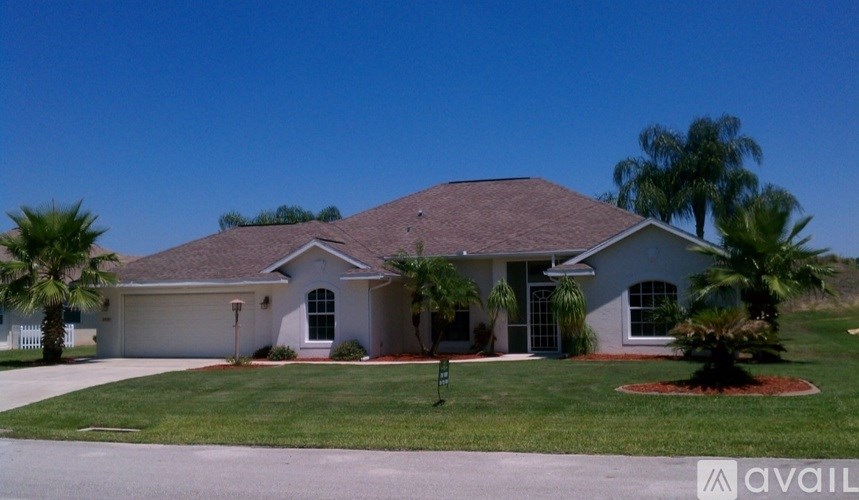 A house with a brown roof and a white exterior is surrounded by a green lawn and palm trees.